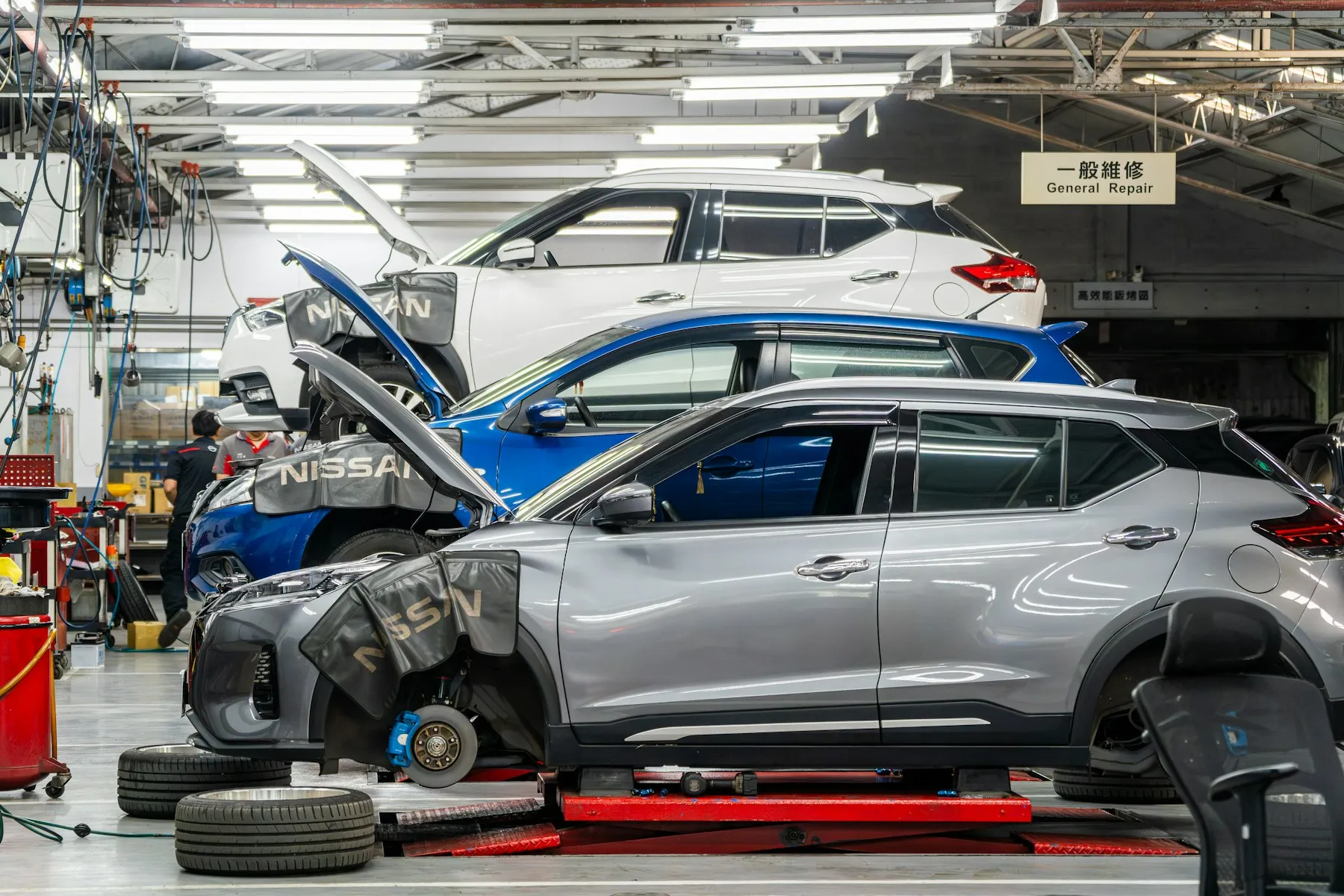 Cars mid-repair in a bright collision repair shop, a silver SUV with its front bumper removed